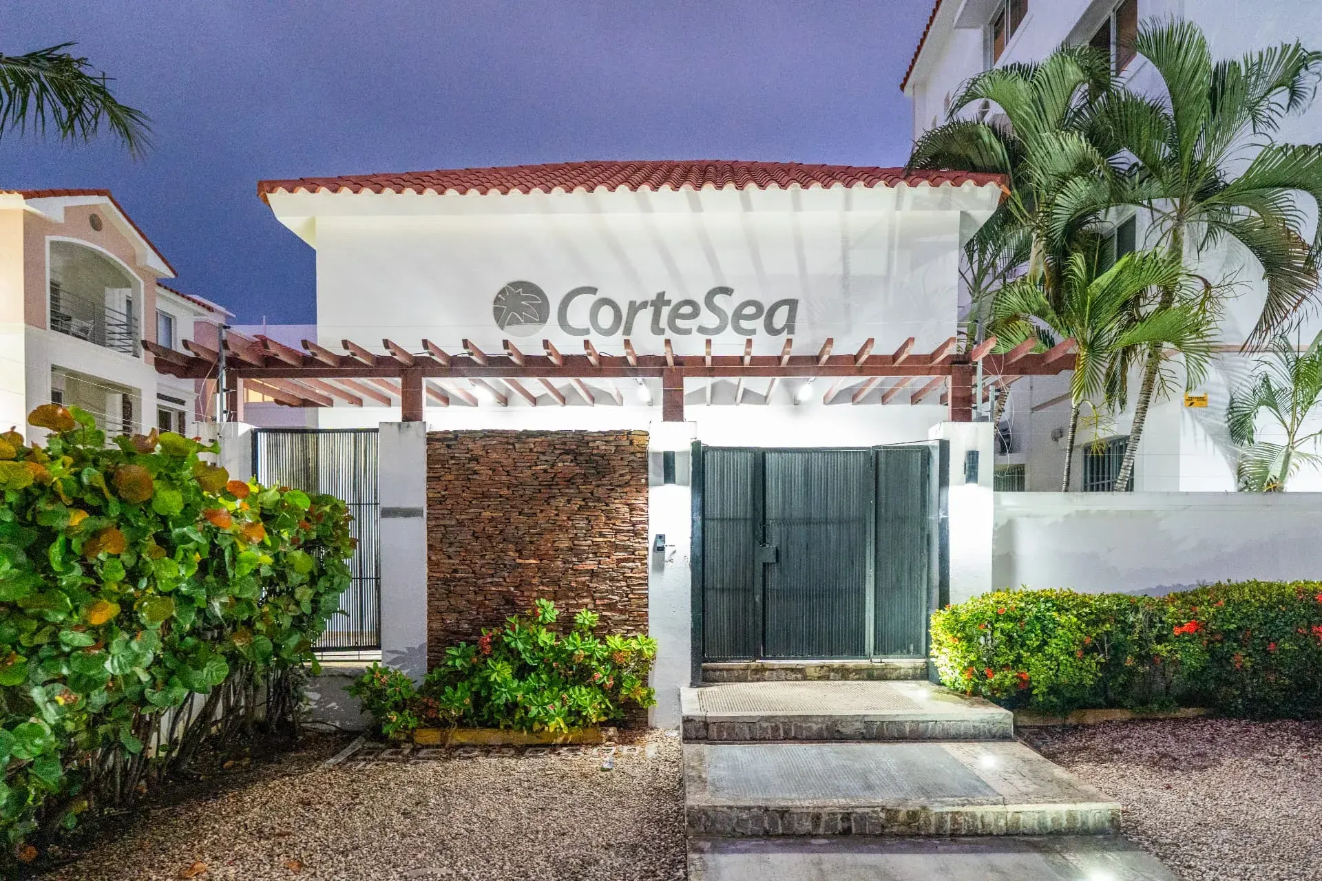 CorteSea gated entrance at dusk with timber pergola, stacked stone wall, uplit palms and flowering hedges
