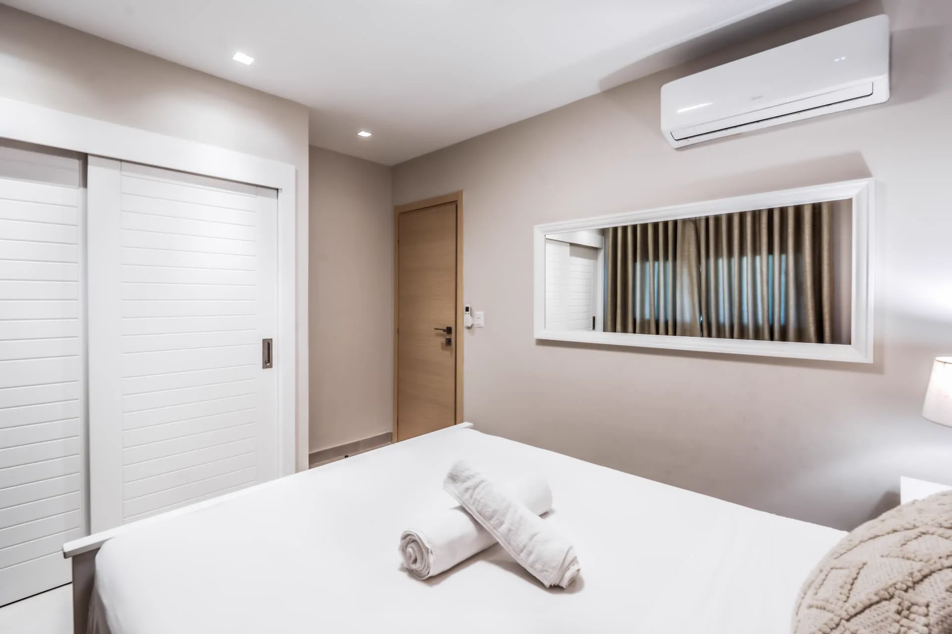 Bedroom with louvred sliding closet doors, framed wall mirror and rolled towels on the bed
