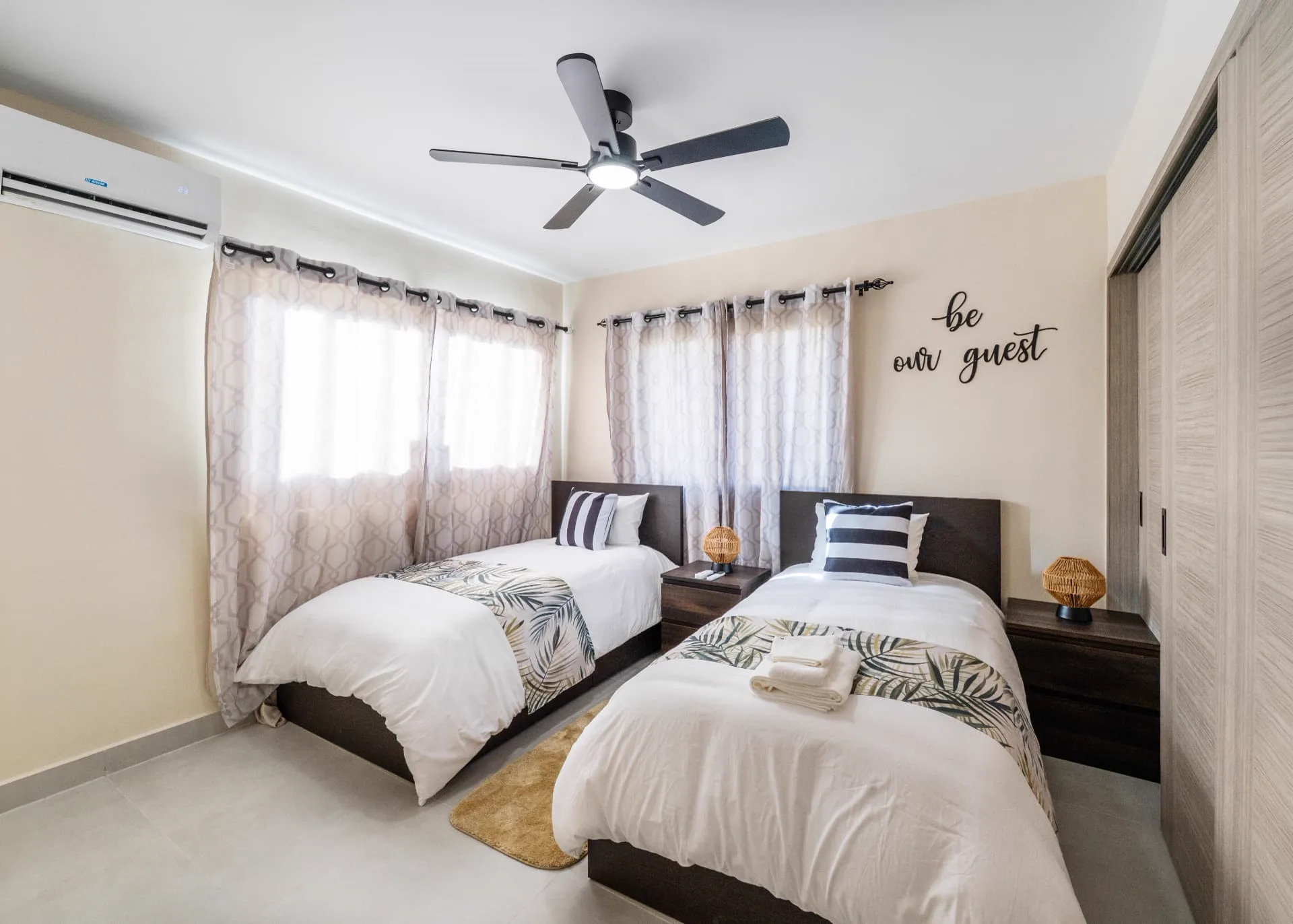 Twin bedroom with two beds, palm-print runners, rattan lamps and a be our guest wall sign