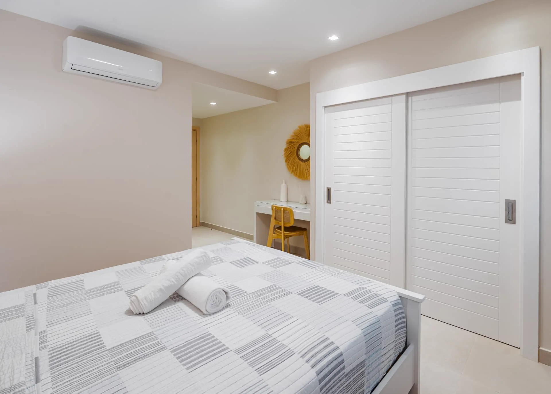 Bedroom with striped coverlet, sliding louvred wardrobe and a compact desk with a woven sunburst mirror