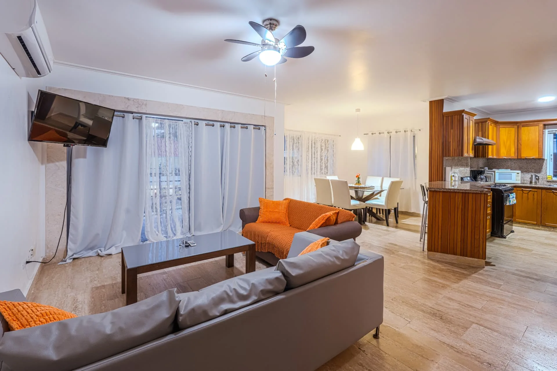 Wide view across the open living room, dining table and kitchen bar with travertine floors and warm timber cabinetry