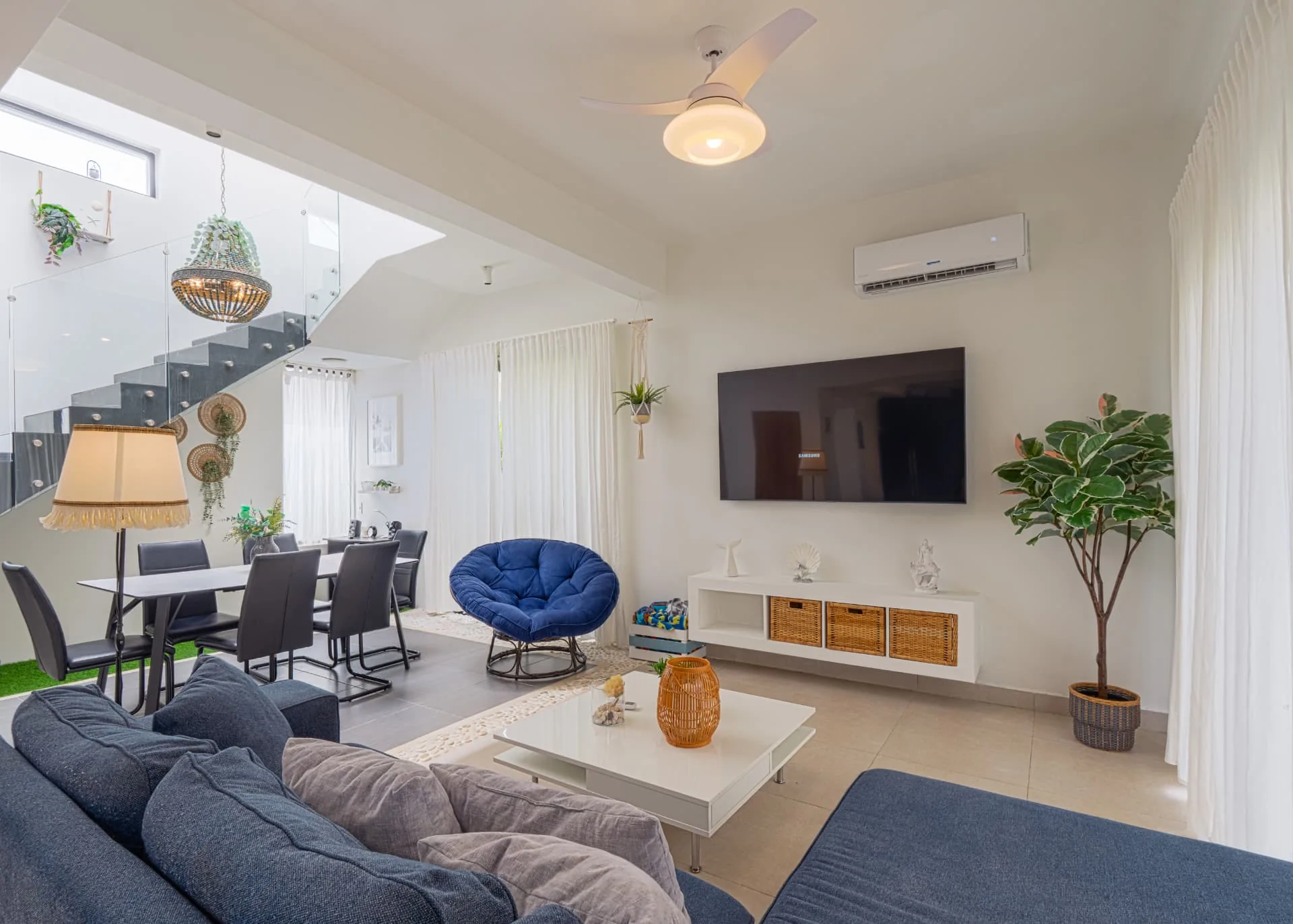 Double-height villa living room with glass staircase, dining table and blue sectional sofa