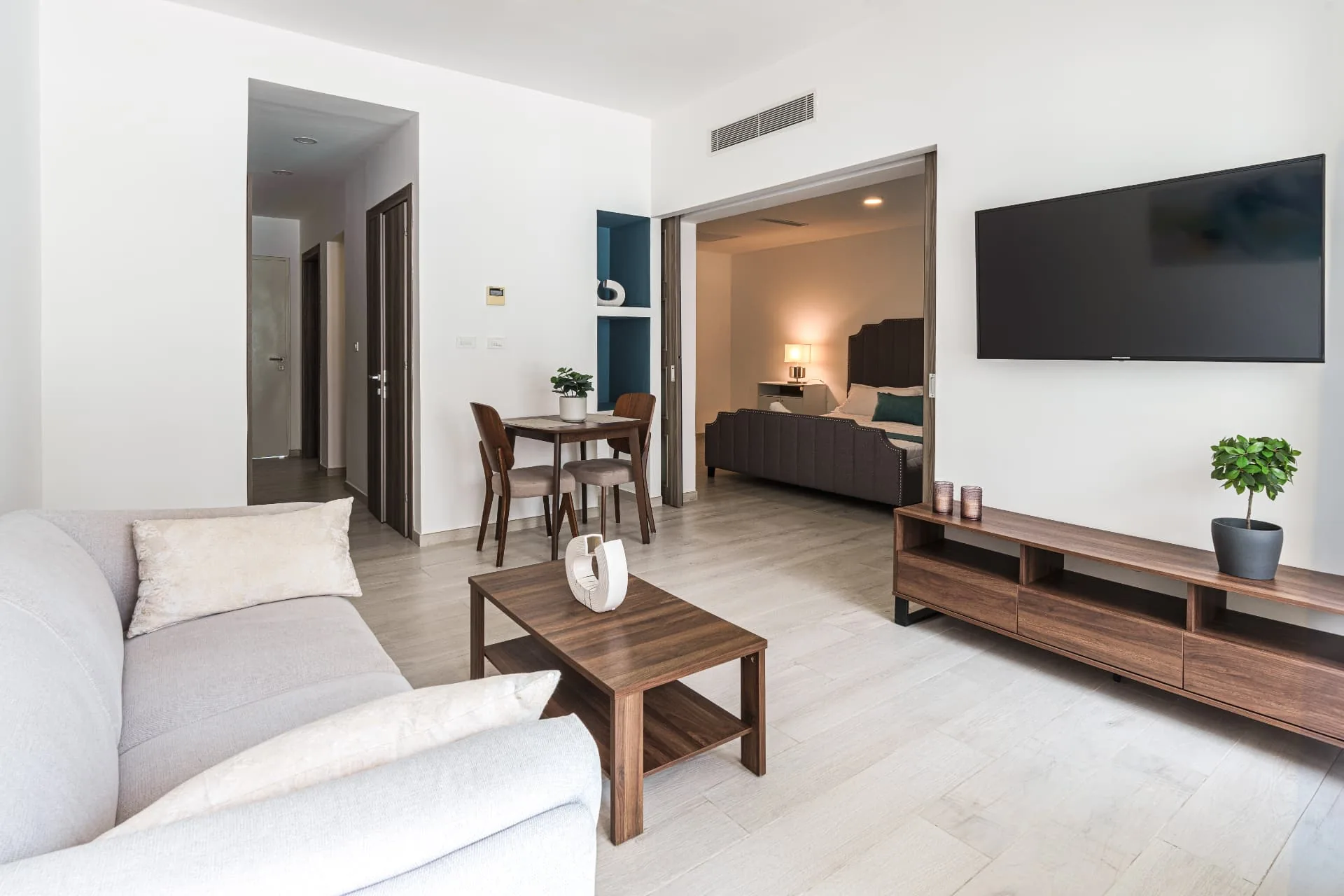 Furnished condominium living room with linen sofa, walnut media console and open bedroom beyond