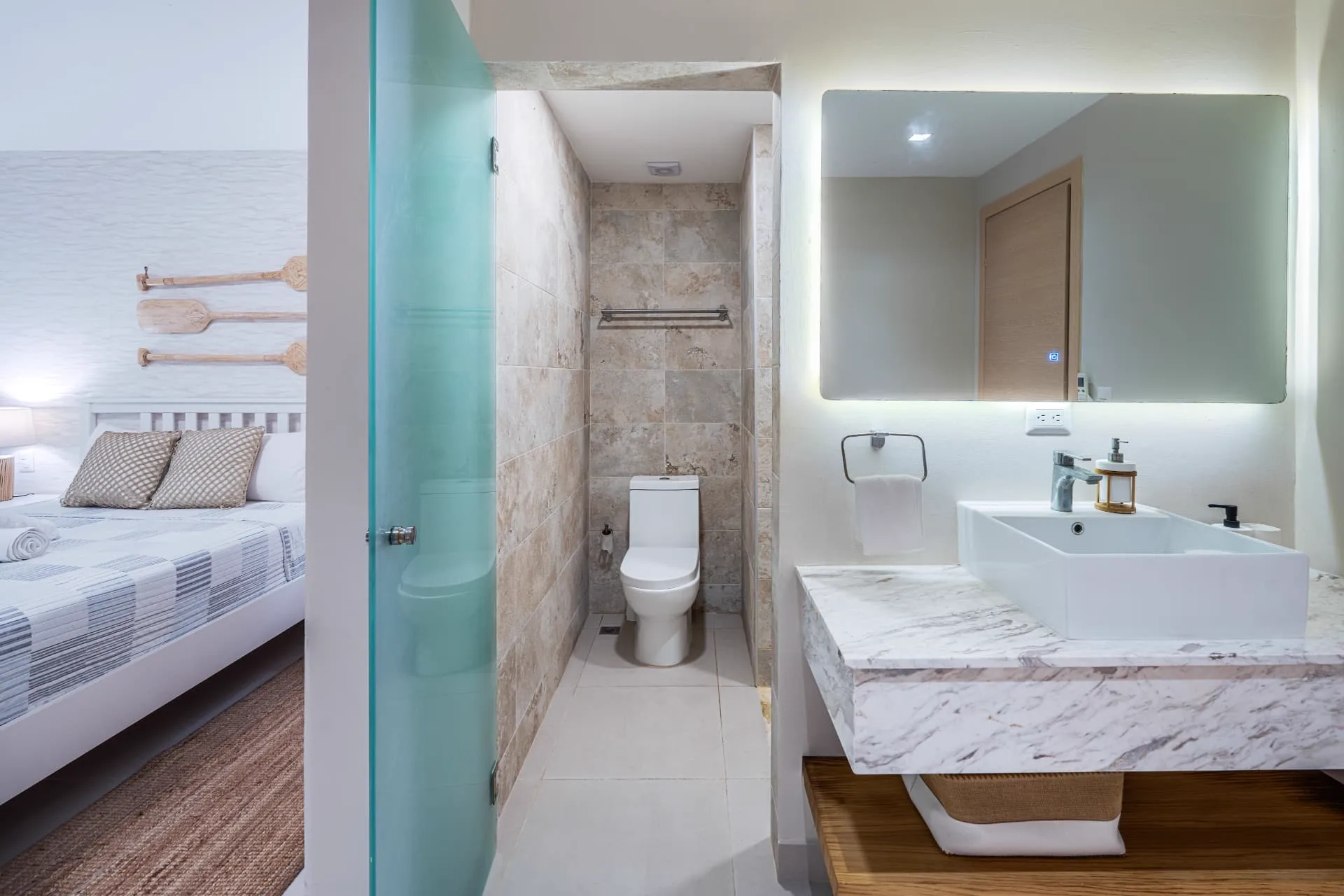 Travertine walk-in shower and toilet behind frosted glass with a marble vanity and backlit mirror