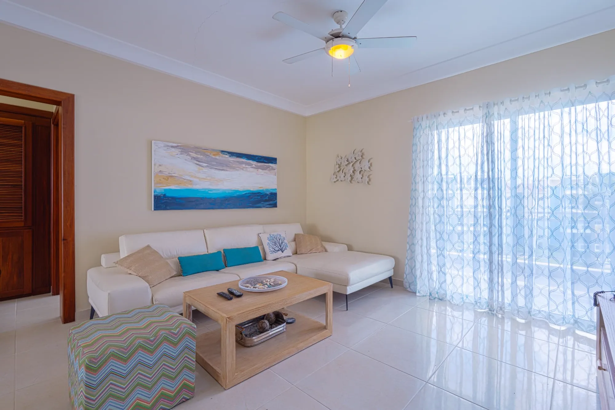 Living room with white leather sectional, turquoise cushions, timber coffee table and full-height sheer curtains