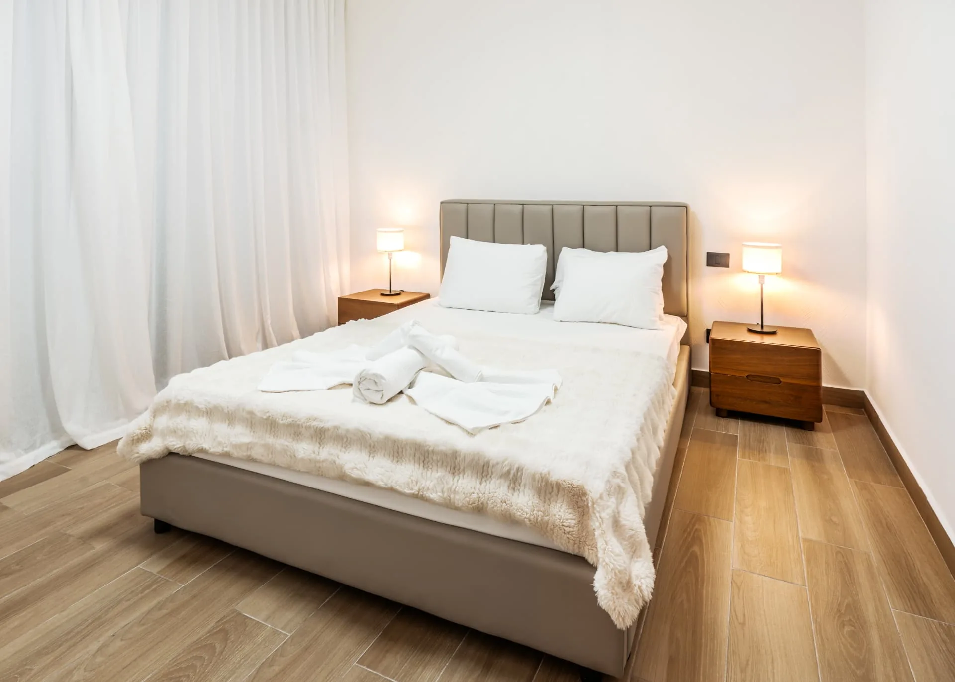 Primary bedroom with tufted headboard, white linens, faux fur throw and matching timber nightstands