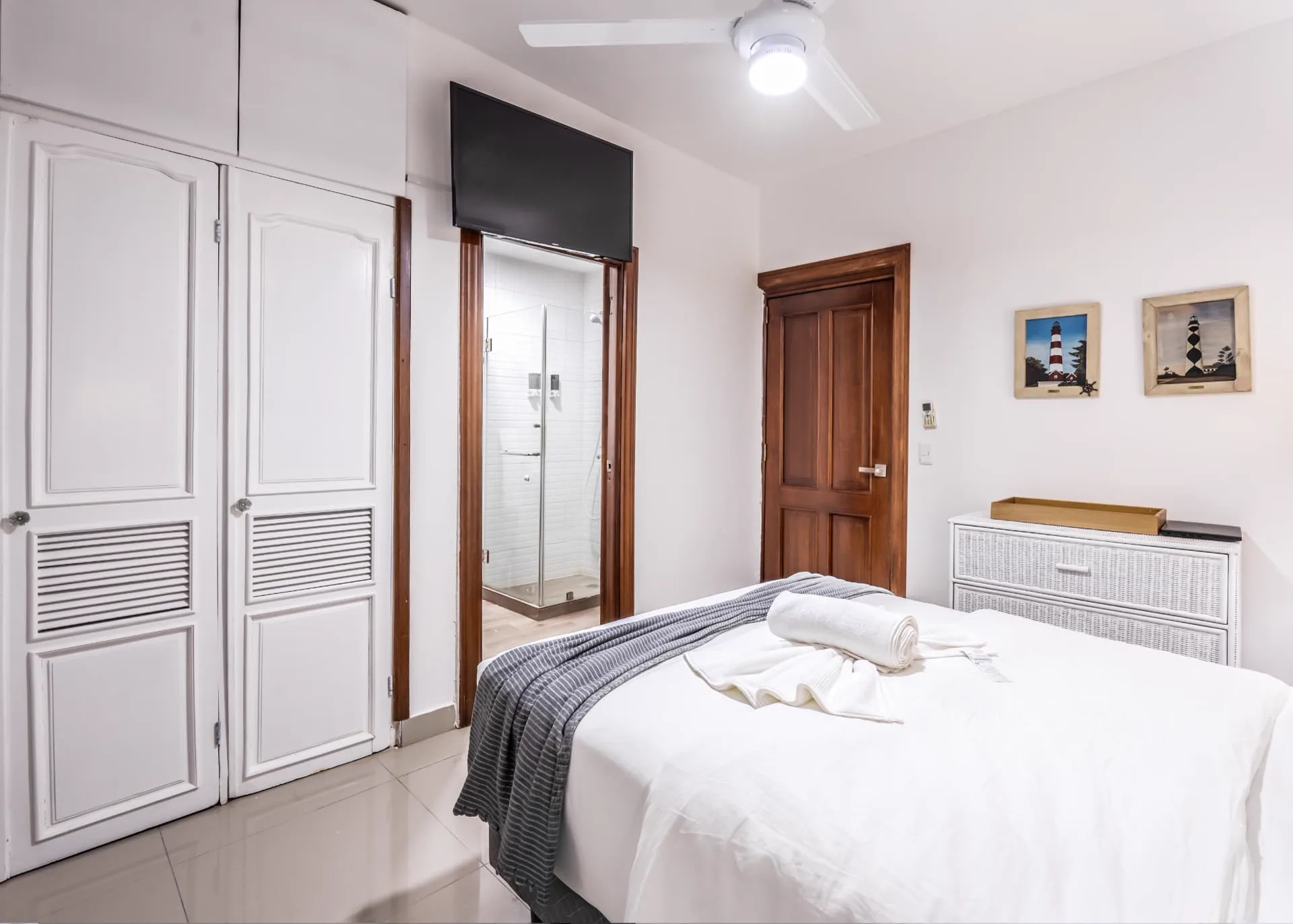 Guest bedroom with wall-mounted television, white louvered wardrobes and an open door onto the glass shower en-suite