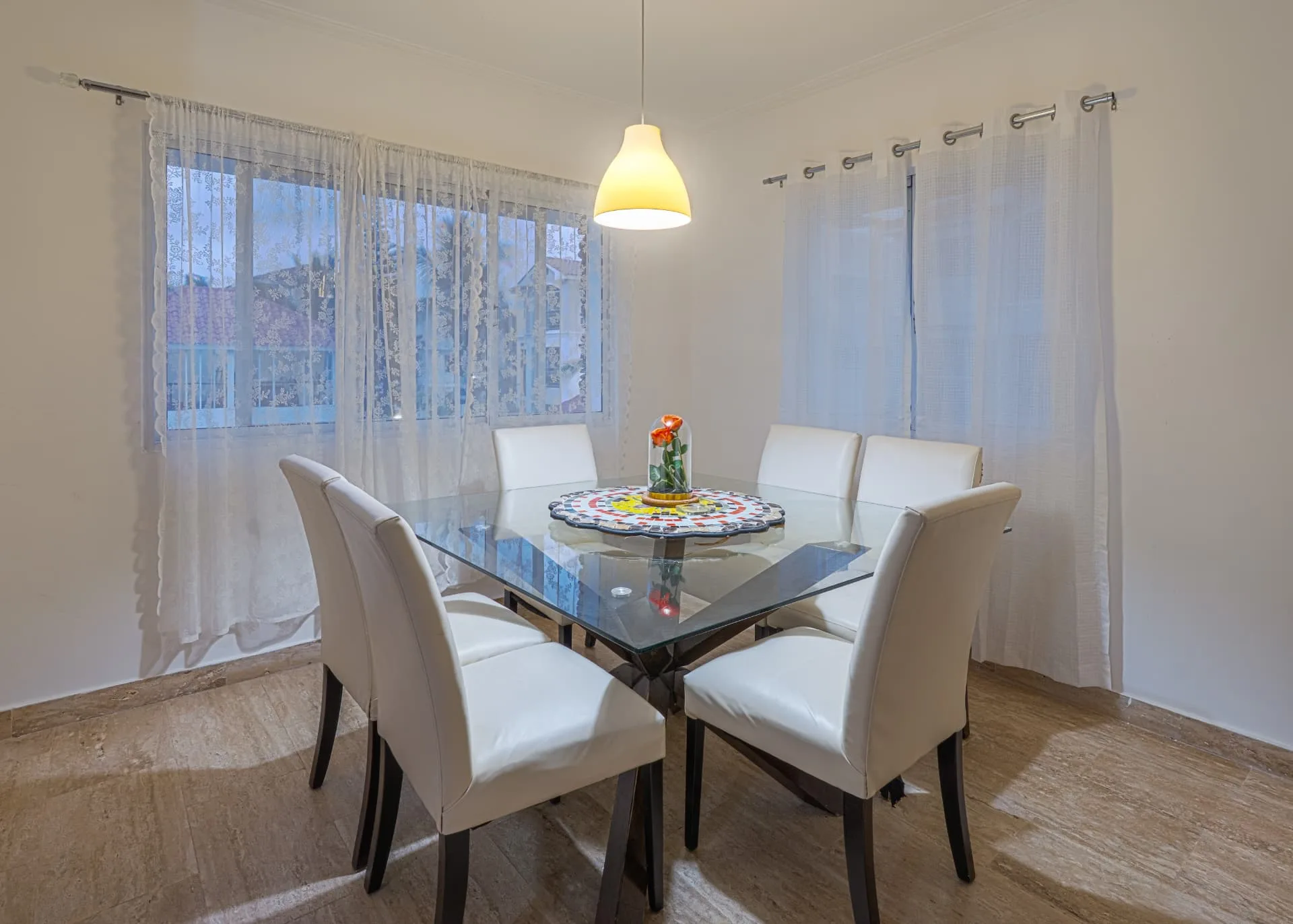 Dining room with glass table, six white leather chairs and a pendant light beside sheer lace curtains