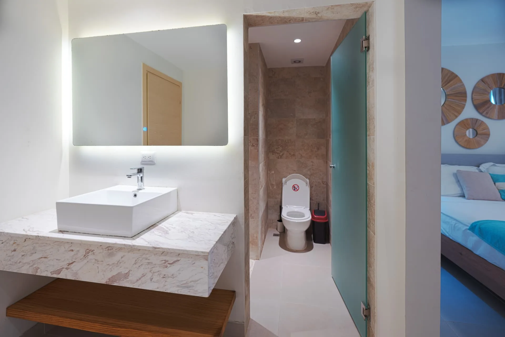 Vanity with vessel sink on a marble counter, backlit mirror and frosted glass door to the washroom
