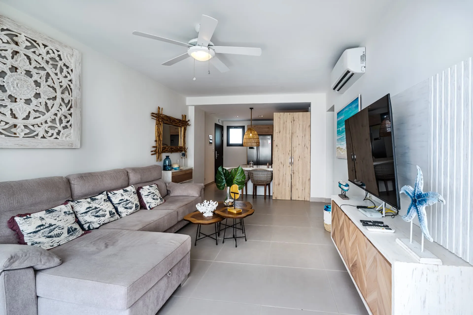 Living room with long grey sectional, carved wood wall art and open sightline through to the kitchen and dining bar