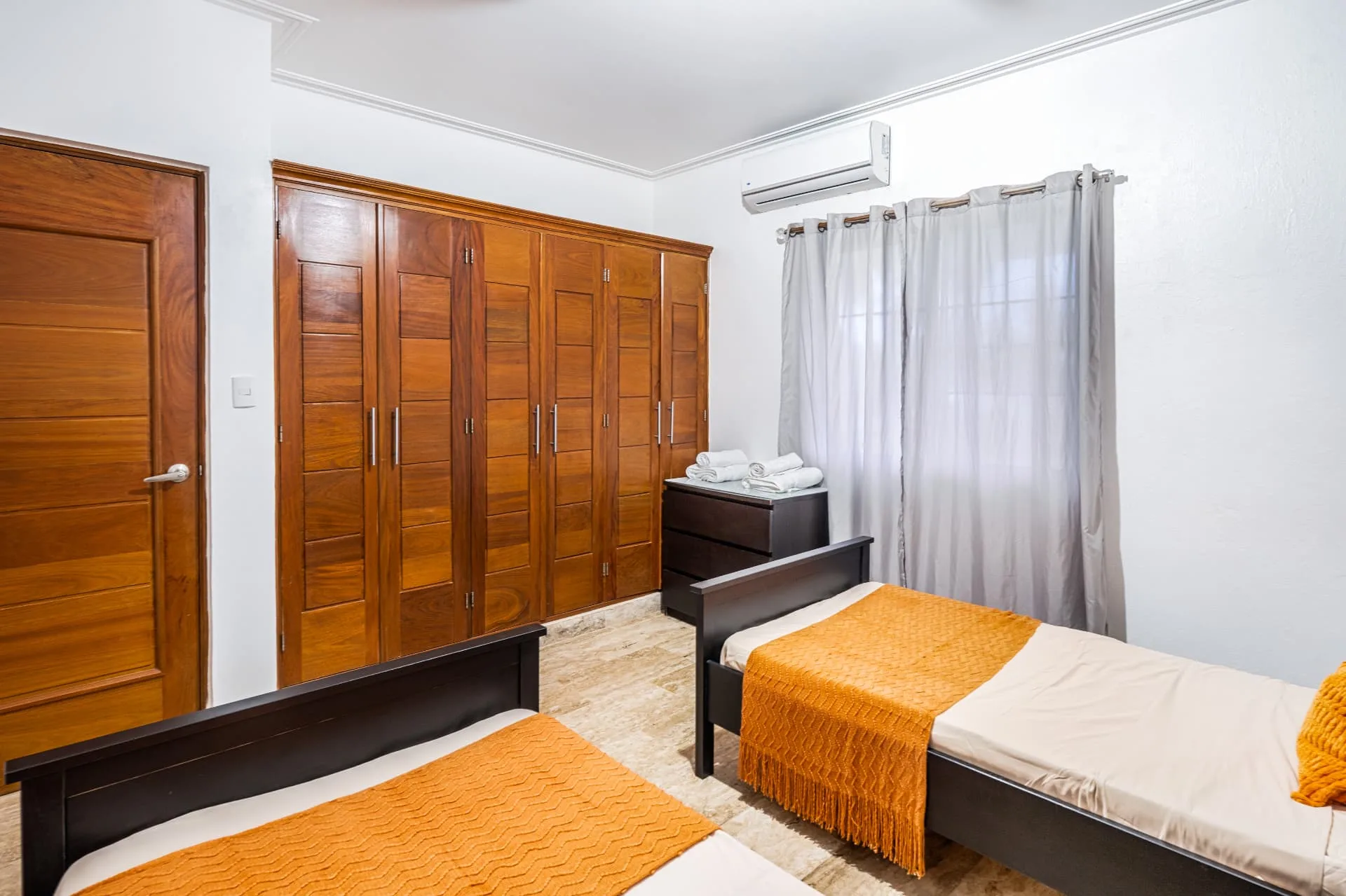 Twin bedroom detail with full-height mahogany closet doors, dresser with folded towels and sheer curtains