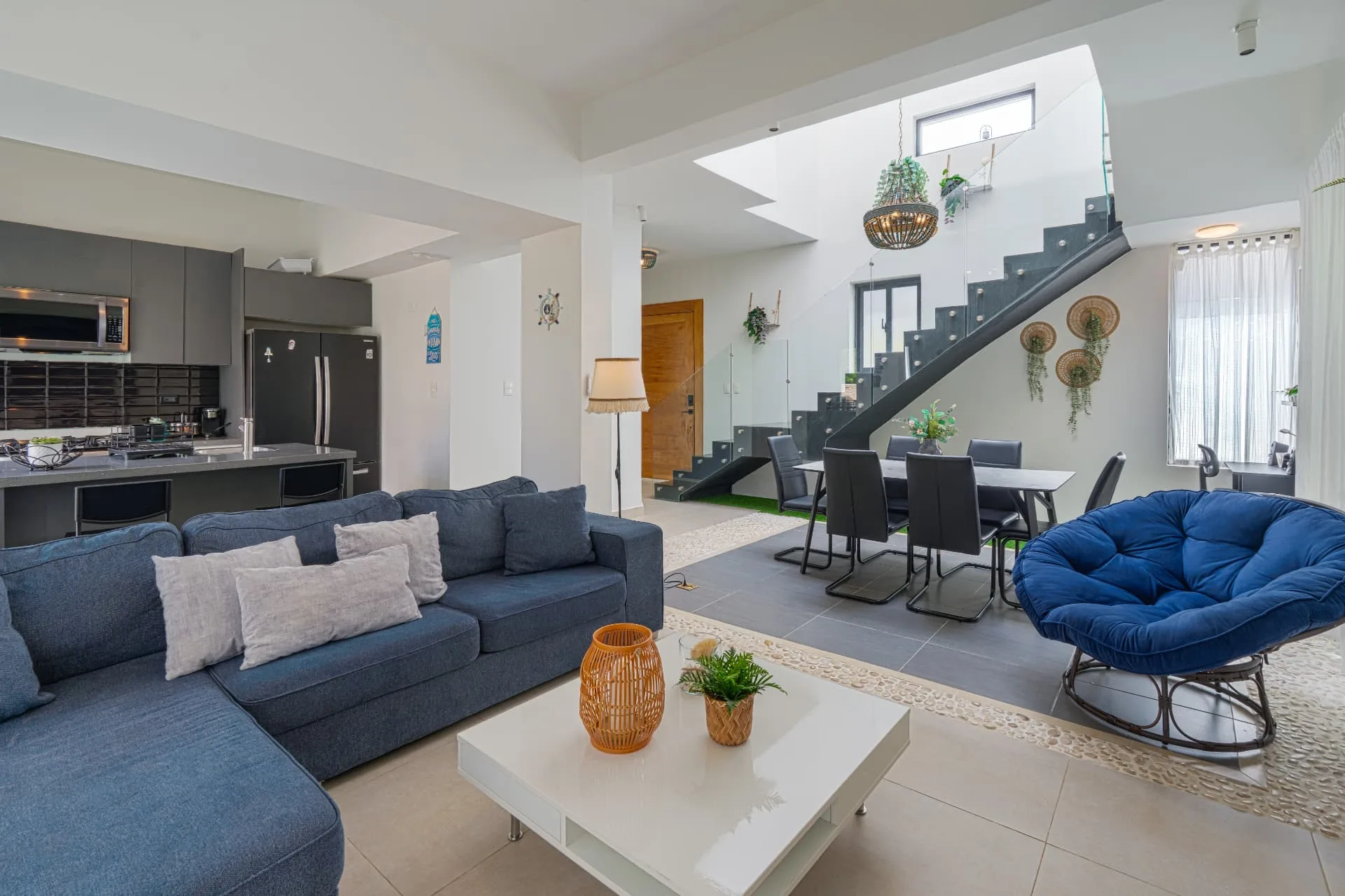 Open double-height living room with blue sectional sofa, glass-railed staircase and dining area