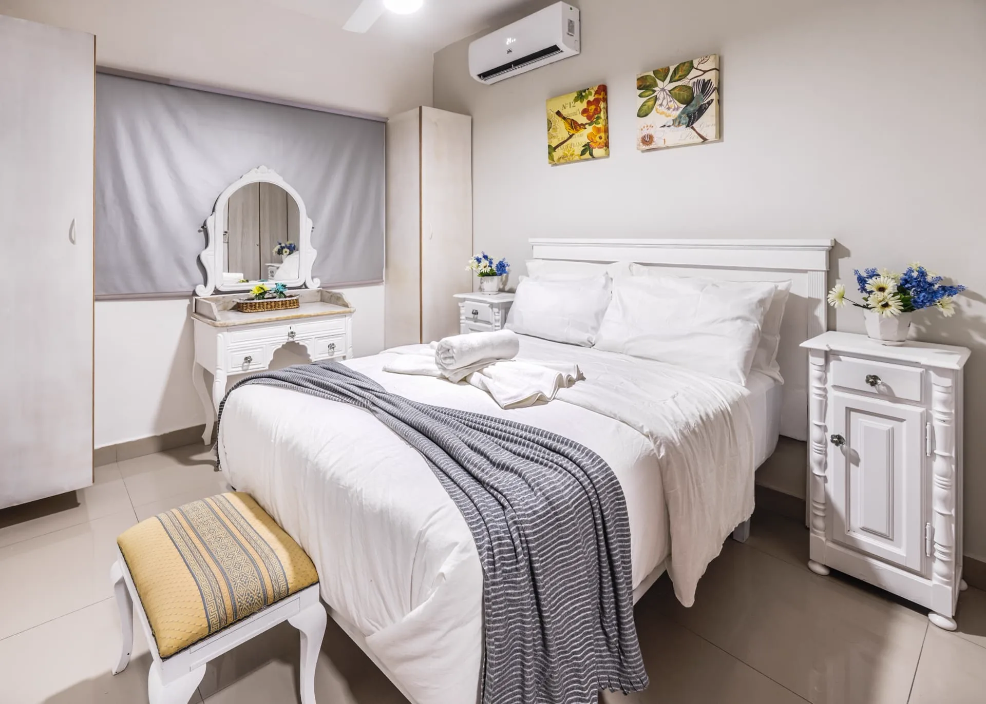 Bedroom with white four-poster bed, grey linen, rolled towels and a blue vase of white flowers