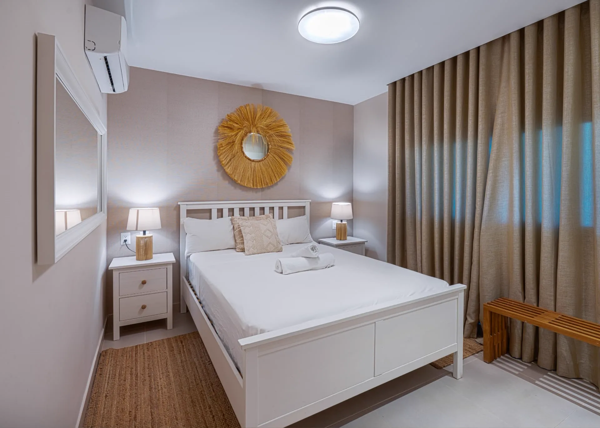 Primary bedroom with white bed frame, woven sunburst mirror, rattan lamps and linen curtains