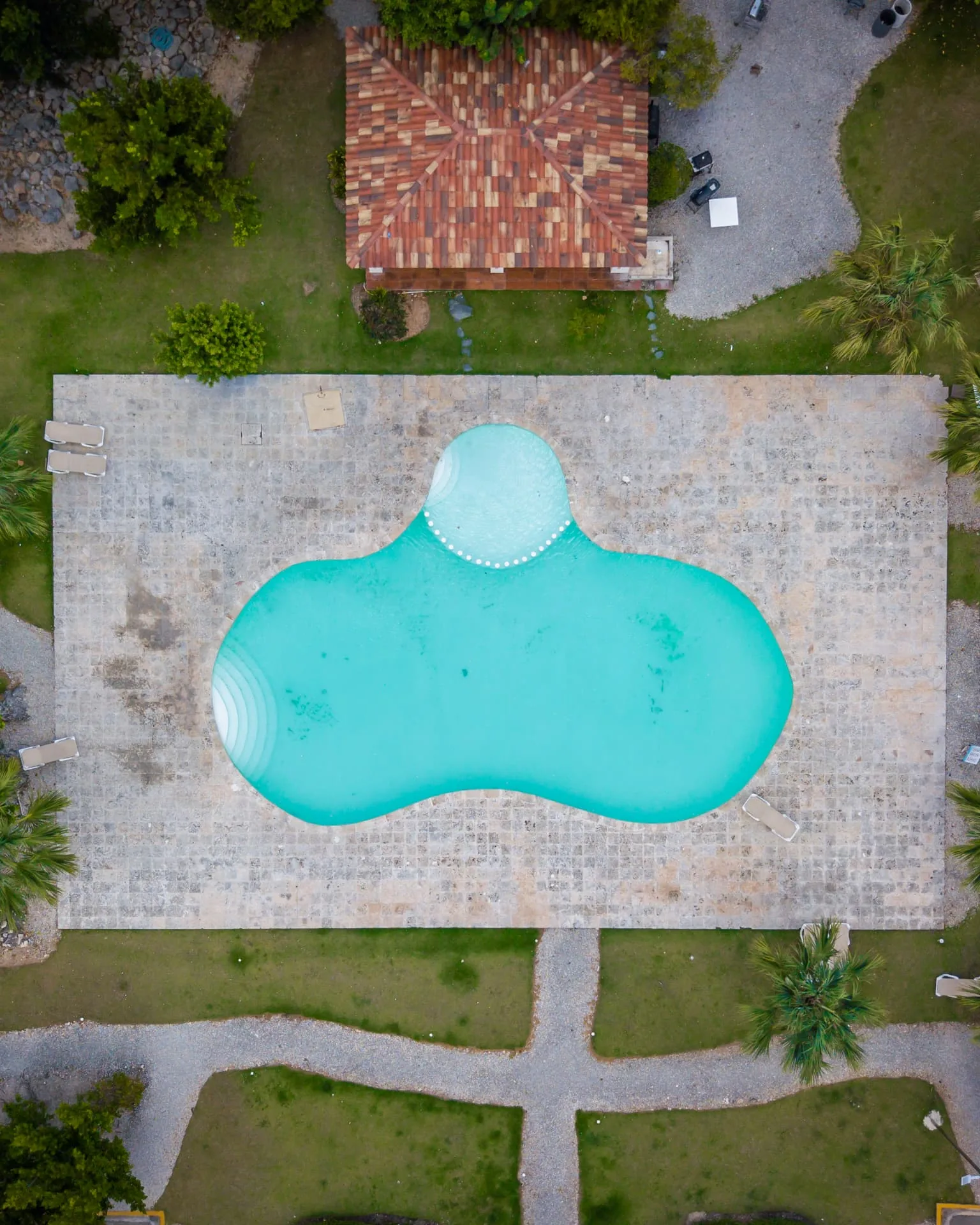 Direct overhead drone view of a free-form turquoise pool with stone deck and thatched pavilion