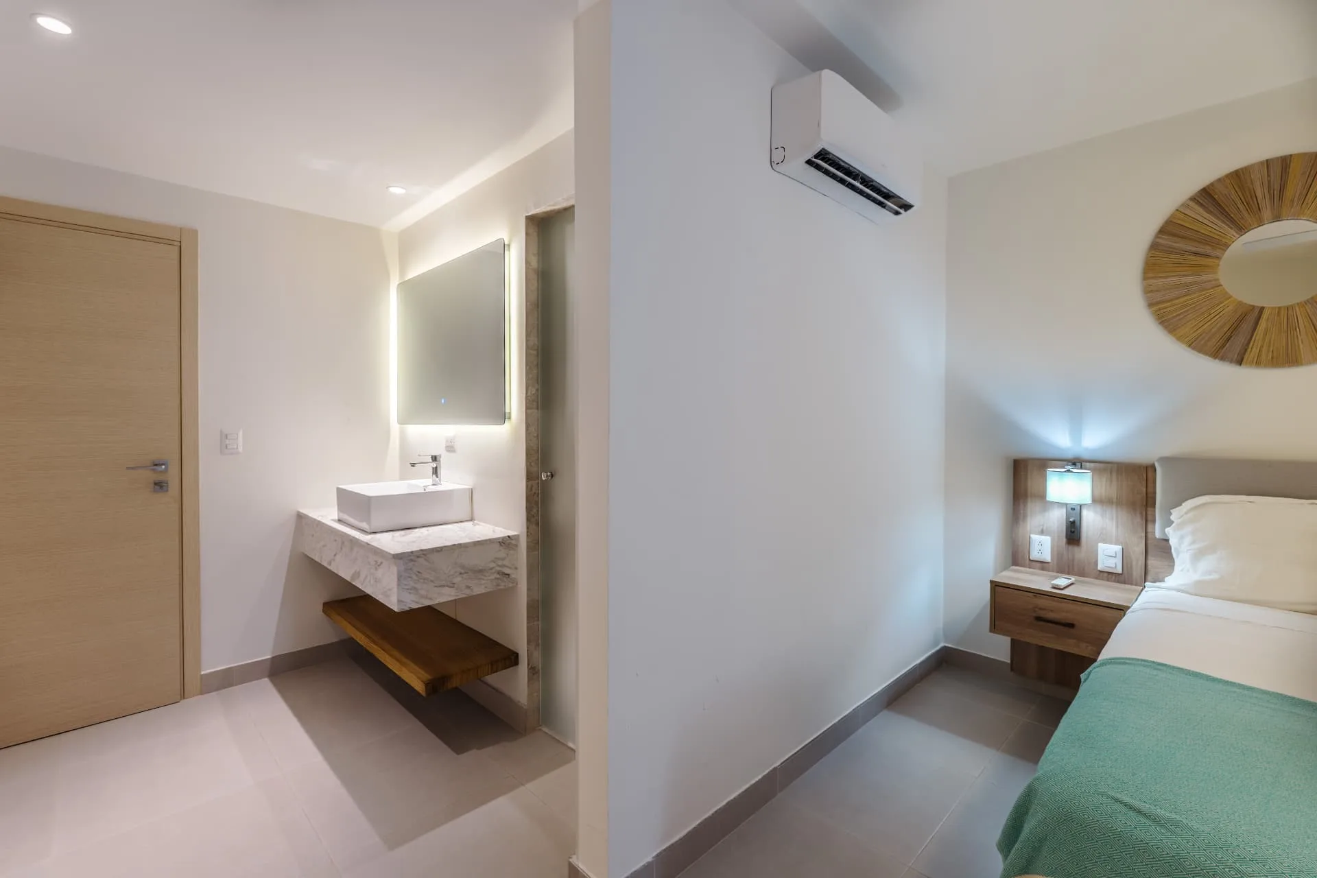 Bedroom with adjoining vanity area, backlit mirror, marble counter and woven sunburst mirror above the bed