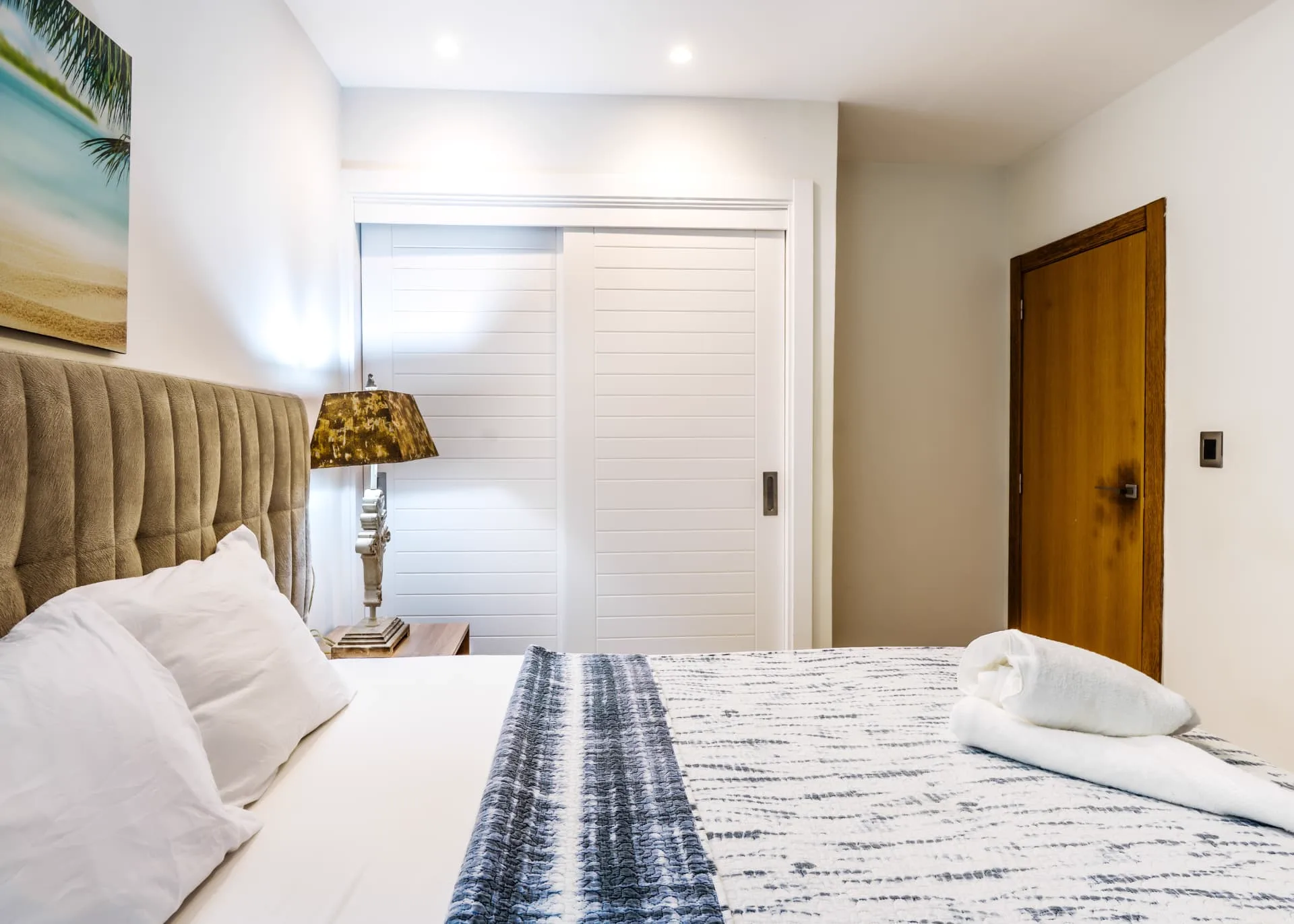 Bedroom detail showing louvred sliding closet doors, bedside lamp and folded towels on the bed