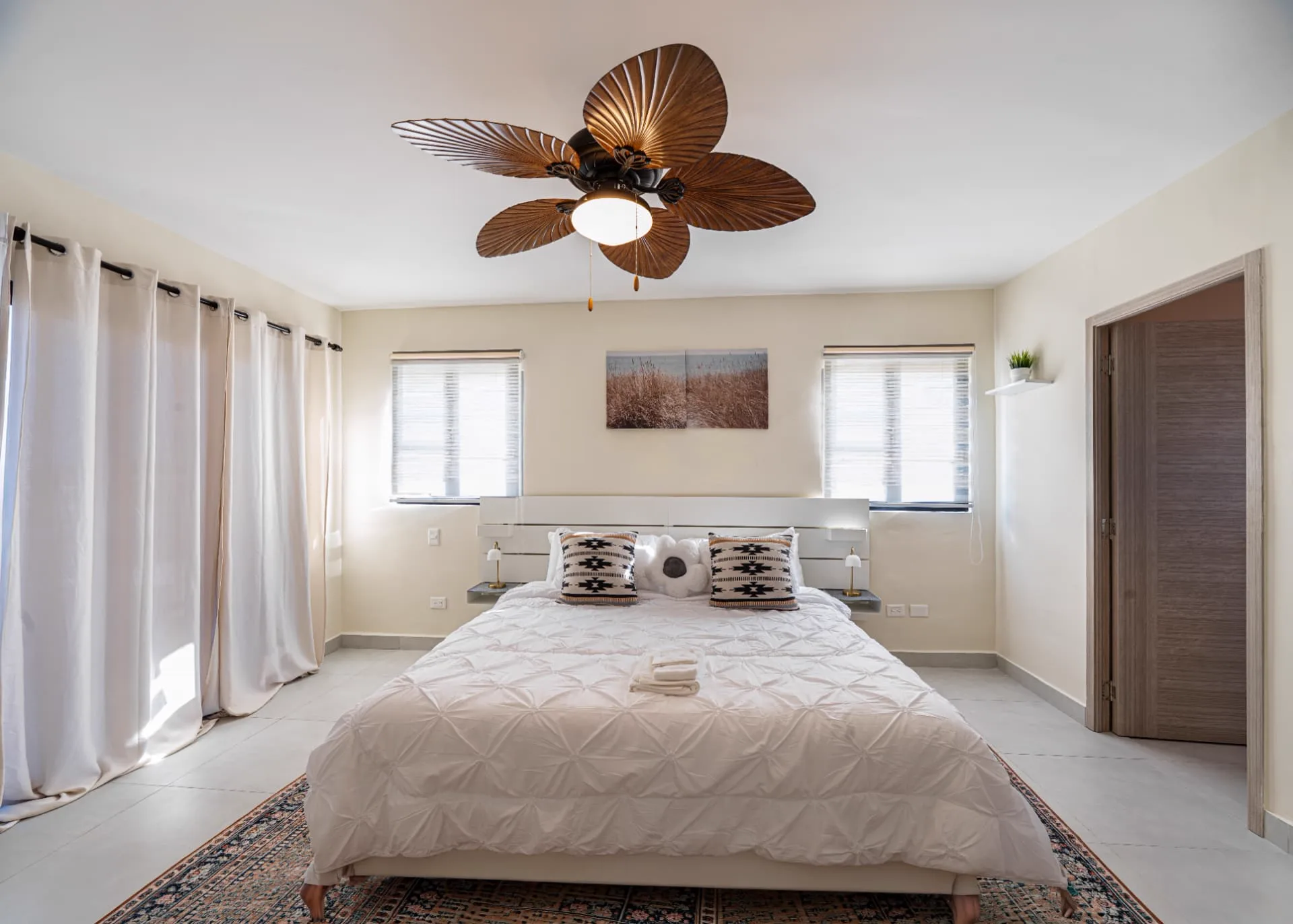 Primary bedroom with king bed, white headboard, palm-leaf ceiling fan and floor-length curtains
