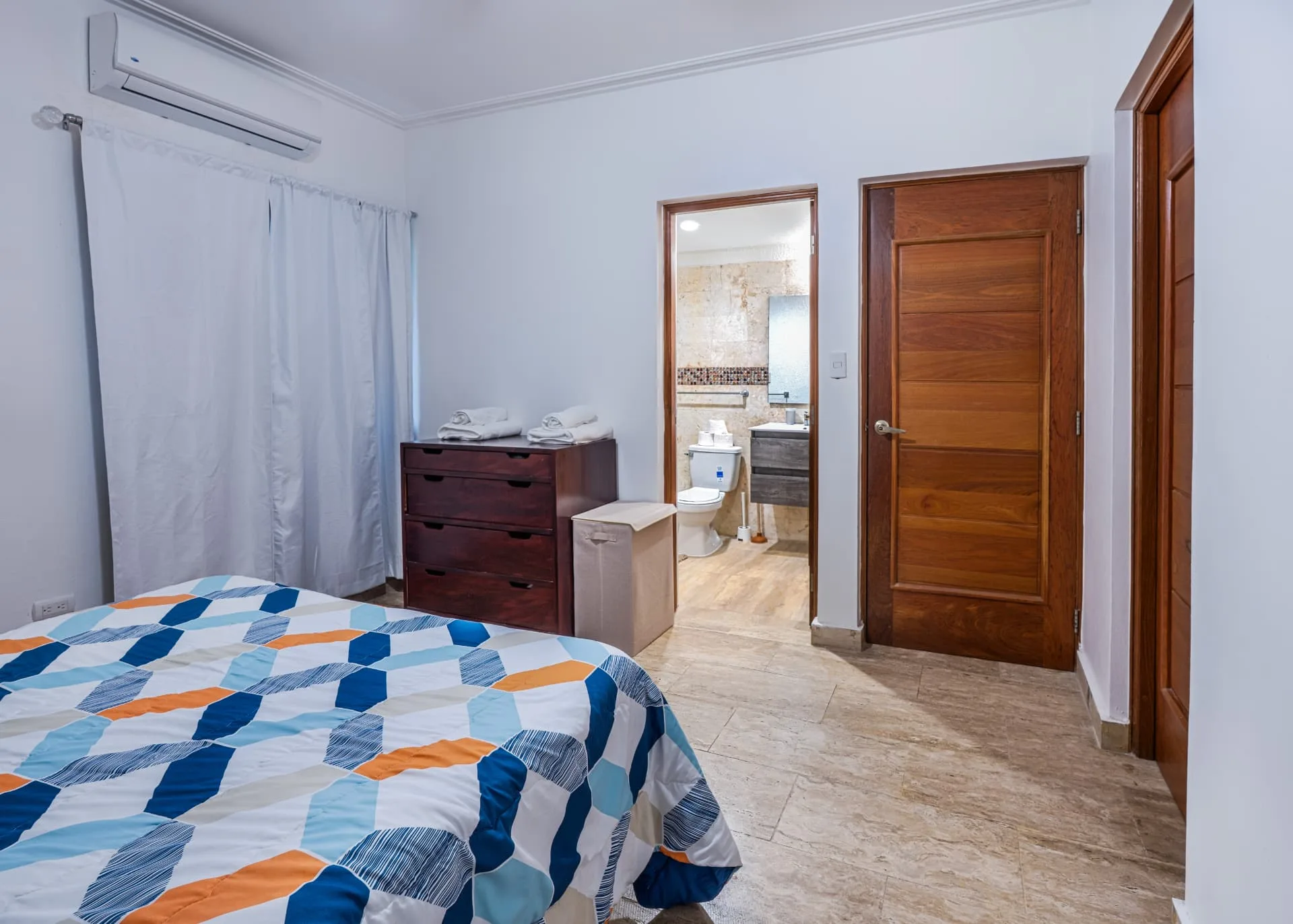 Bedroom view towards the en-suite bathroom with a mahogany dresser, folded towels and travertine floors
