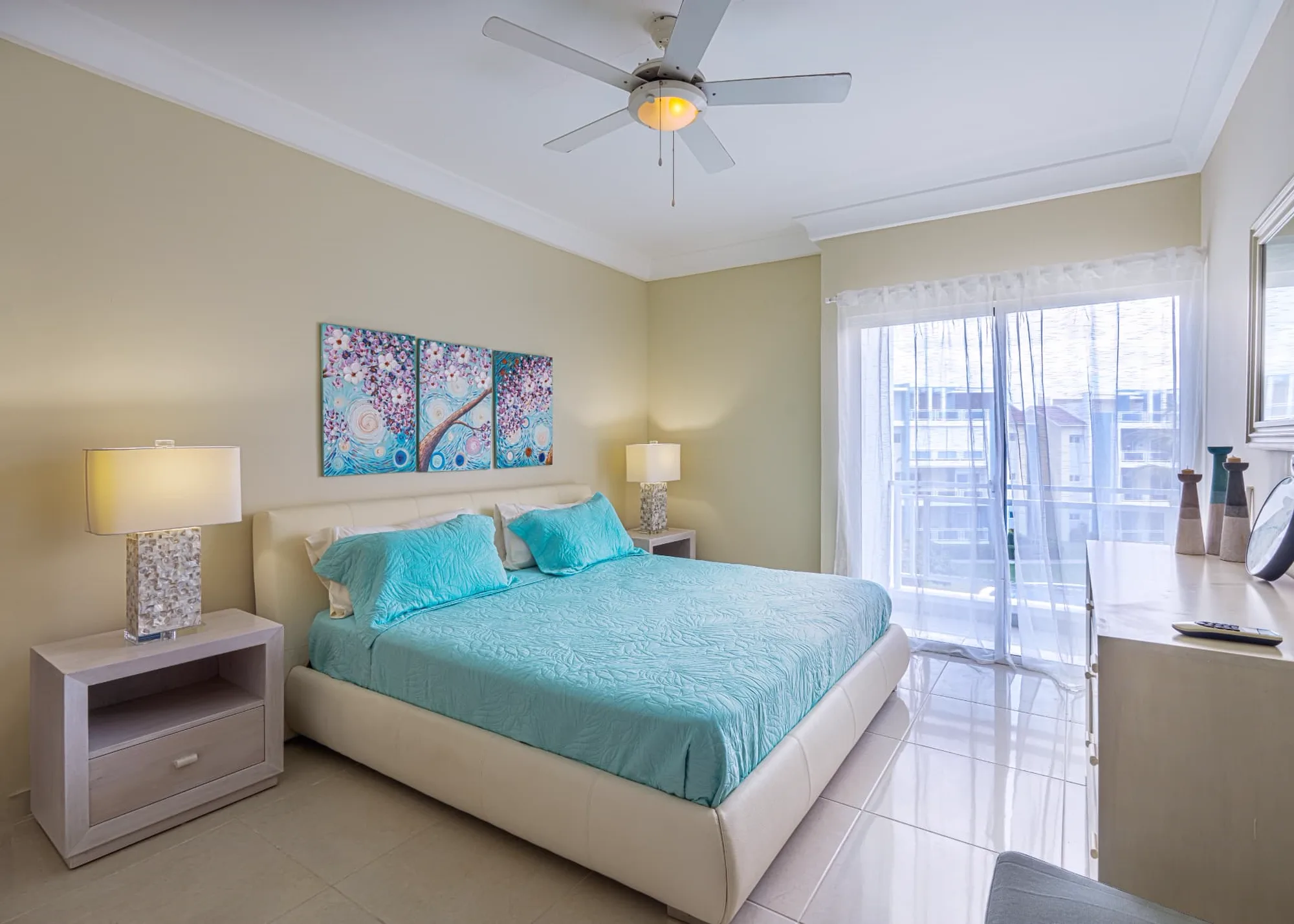 Primary bedroom with king bed, turquoise quilt, triptych blossom artwork and balcony doors behind sheer curtains